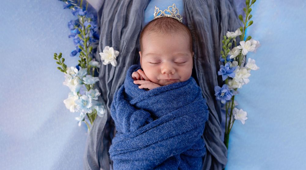 Newborn baby swaddled in blue wrap with floral bow headband, sleeping peacefully among white and blue flowers.