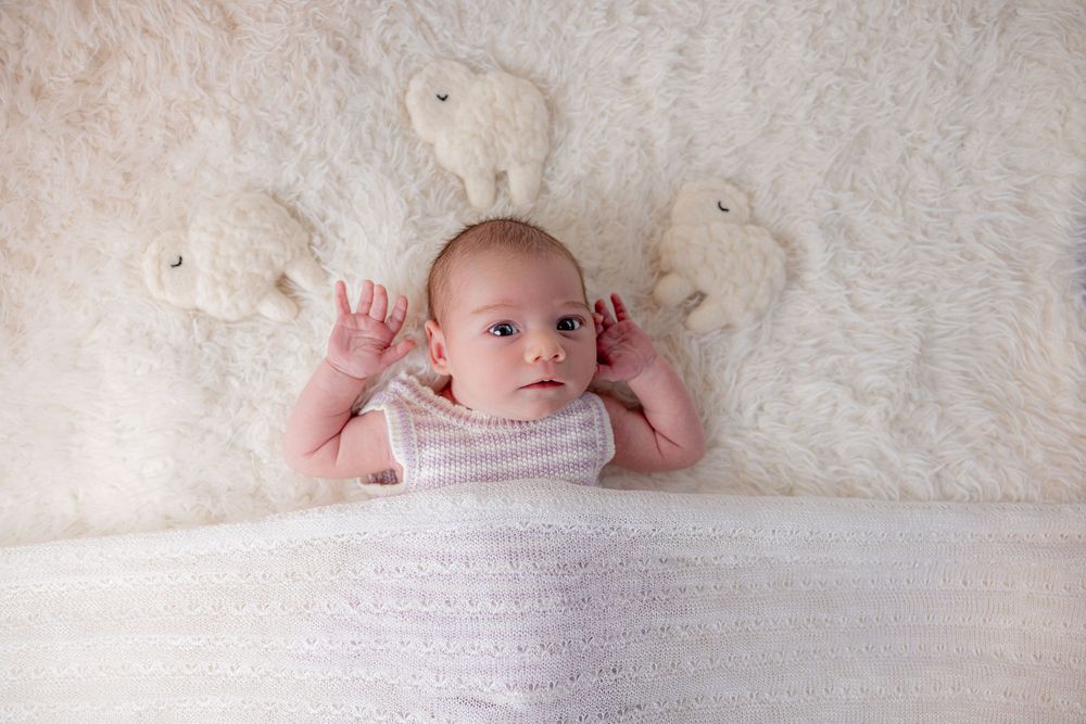 Adorable baby lying on fluffy white fur blanket surrounded by soft elephant stuffed animals in a cozy nursery setting.