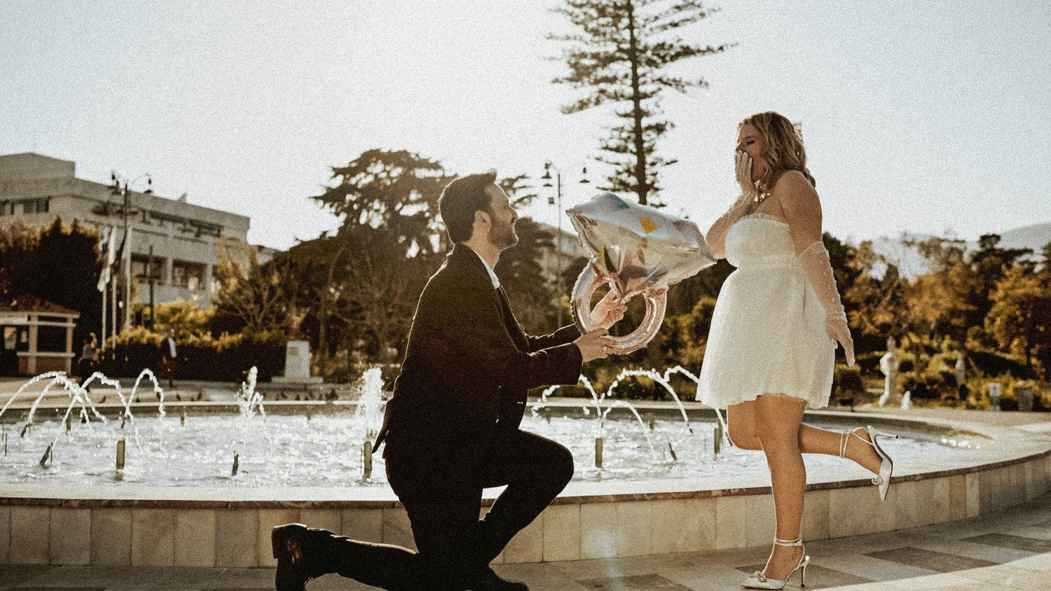 A romantic proposal scene at sunset with a couple by a fountain in a park setting.