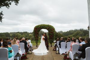 Outdoor wedding ceremony with bride and groom at floral arch, guests seated on white chairs, lush green trees backdrop.