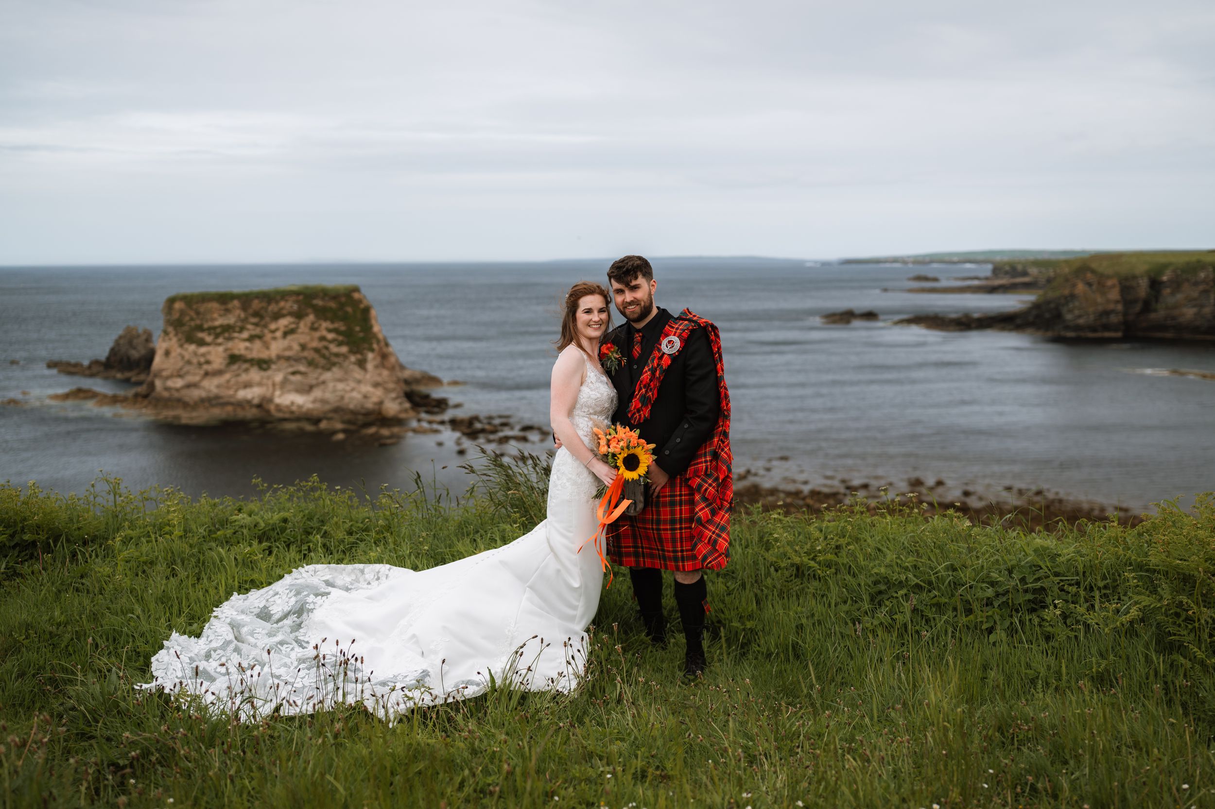 Bride and groom in Scottish kilt pose on coastal cliffs with sunflowers, sea stacks, and ocean views on their wedding day.