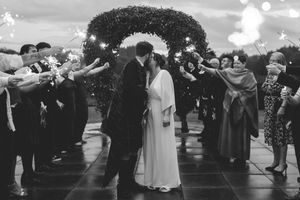 Black and white photo of newlyweds kissing under sparklers held by wedding guests outdoors.