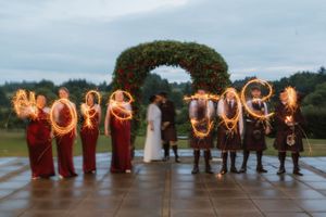 Wedding party holding sparklers spelling Mr & Mrs under floral arch at twilight outdoor ceremony.