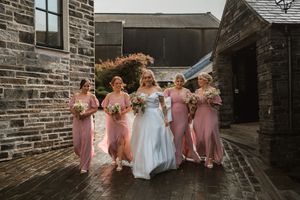 Bride in white gown walks with four bridesmaids in pink dresses holding bouquets outside a stone venue.