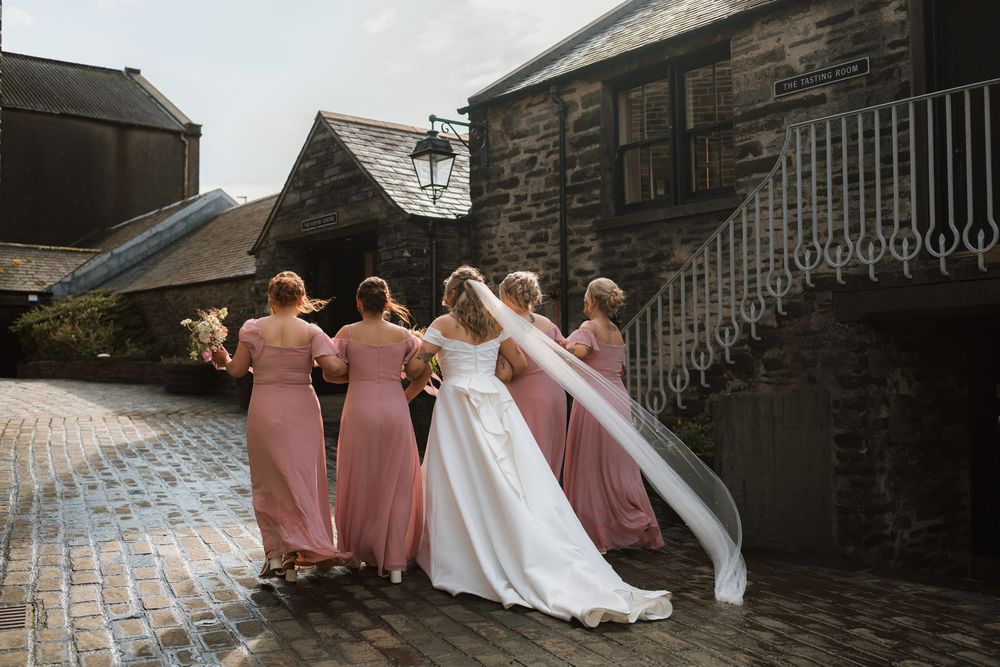 Bride in white gown and veil stands with four bridesmaids in pink dresses on cobblestone courtyard outside stone venue.