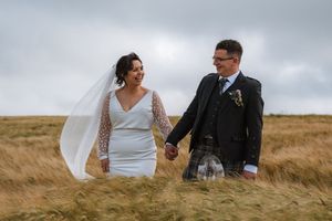 Bride and groom holding hands walking through golden grass fields in Scotland, bride in white gown with veil, groom in tartan kilt.