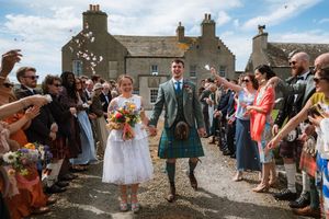 Bride and groom walk through confetti at outdoor Scottish wedding, groom wearing traditional tartan kilt, guests cheering.