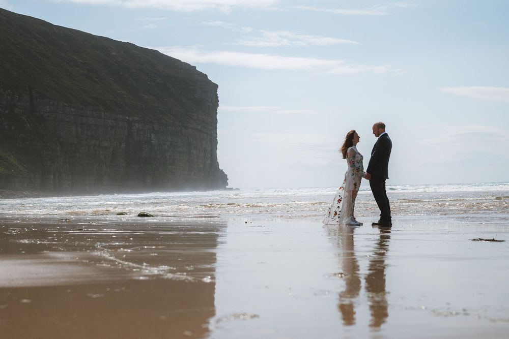Couple in wedding attire holding hands on a reflective sandy beach with dramatic coastal cliffs in the background.