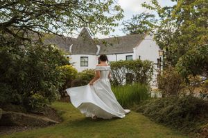 Bride in flowing white gown twirling in lush garden outside a white country house.