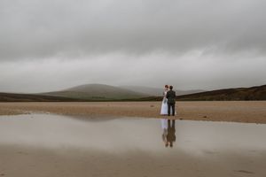 Couple embracing on a vast sandy beach with misty hills and dramatic cloudy sky reflected in shallow water.
