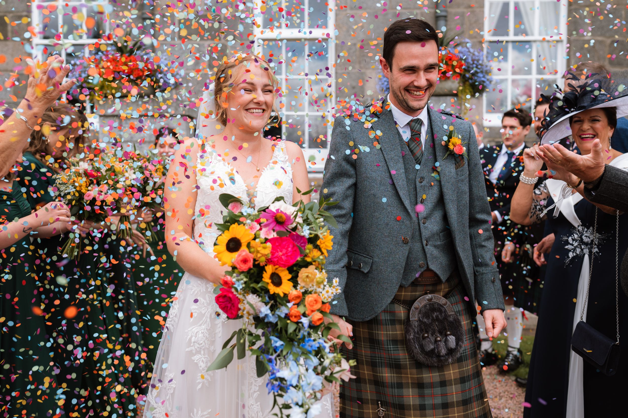Wedding couple celebrates under a shower of colorful confetti while holding vibrant autumn flowers and smiling joyfully.