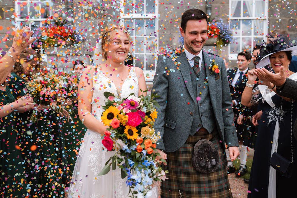 Wedding couple celebrates under a shower of colorful confetti while holding vibrant autumn flowers and smiling joyfully.