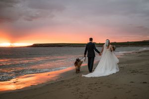 Bride and groom walk hand-in-hand with their dog on a beach at sunset in wedding attire.