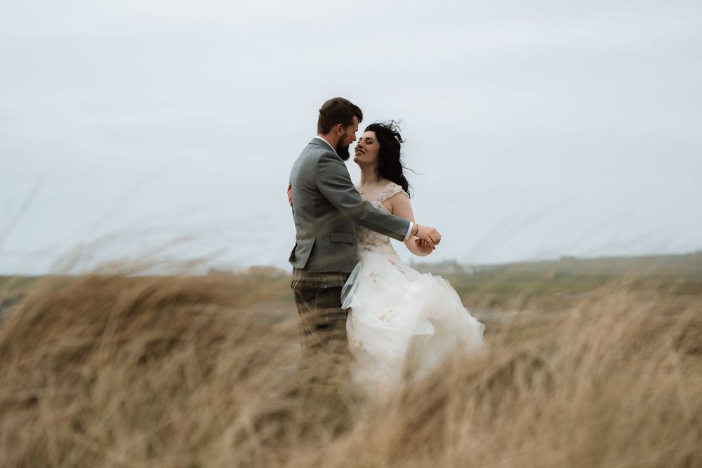 Two figures embrace romantically in a windswept field of tall grass under an overcast sky.