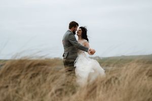 Two figures embrace romantically in a windswept field of tall grass under an overcast sky.