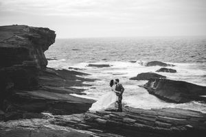 Two silhouettes stand together on dramatic coastal cliffs overlooking rough ocean waves in black and white.