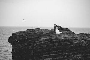 A couple stands on a dramatic rocky cliff overlooking the ocean in a black and white artistic photograph.