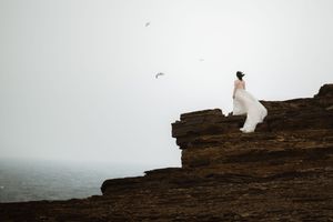 Someone in a white dress stands on dramatic rocky cliffs overlooking a misty ocean landscape with birds flying above.
