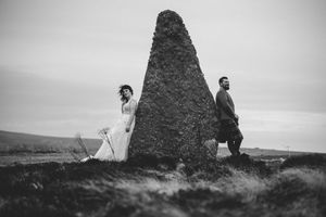A black and white wedding photo showing a couple standing on opposite sides of a large stone monolith in a rural setting.