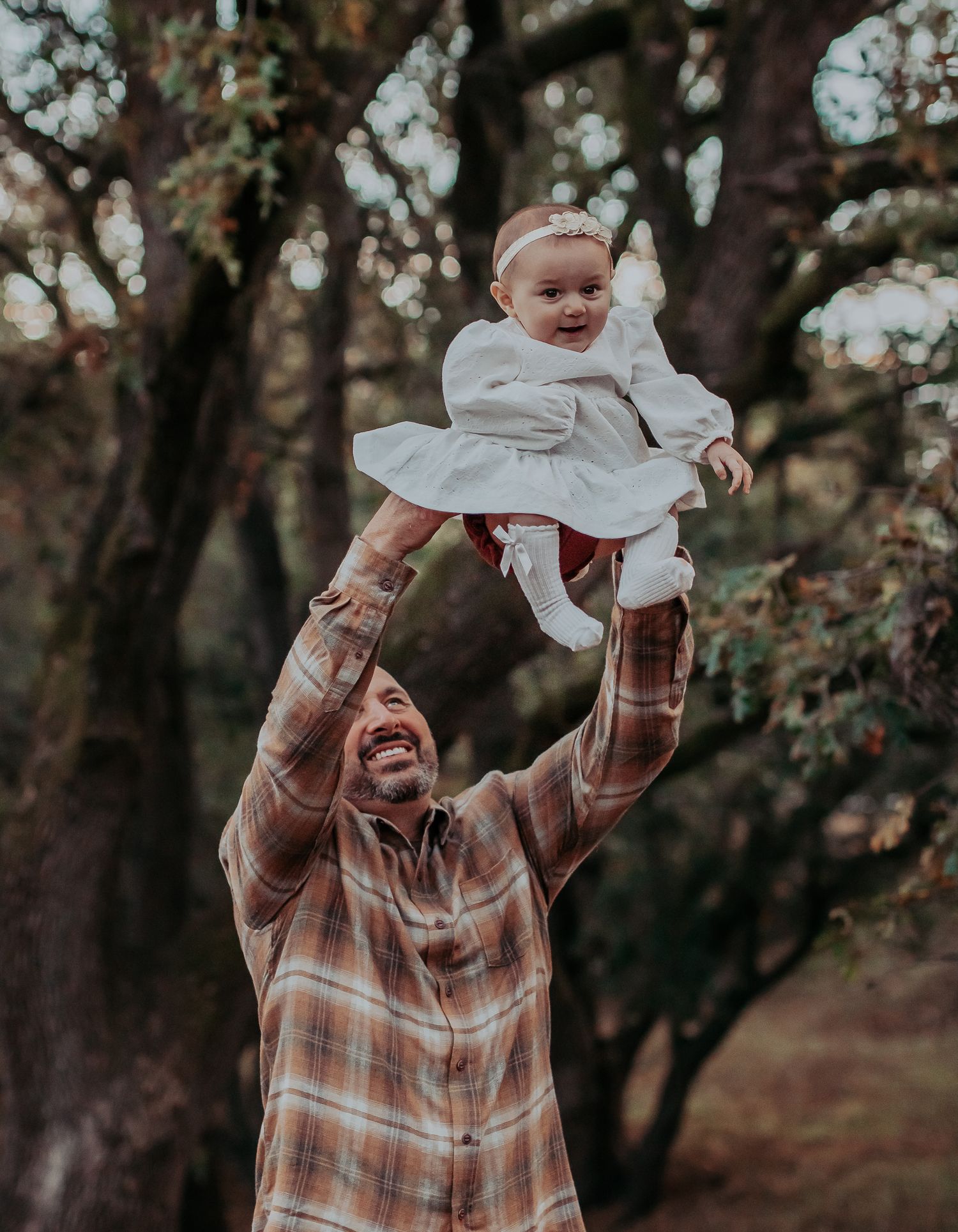 Father in plaid shirt joyfully lifts baby in white dress during outdoor photoshoot.
