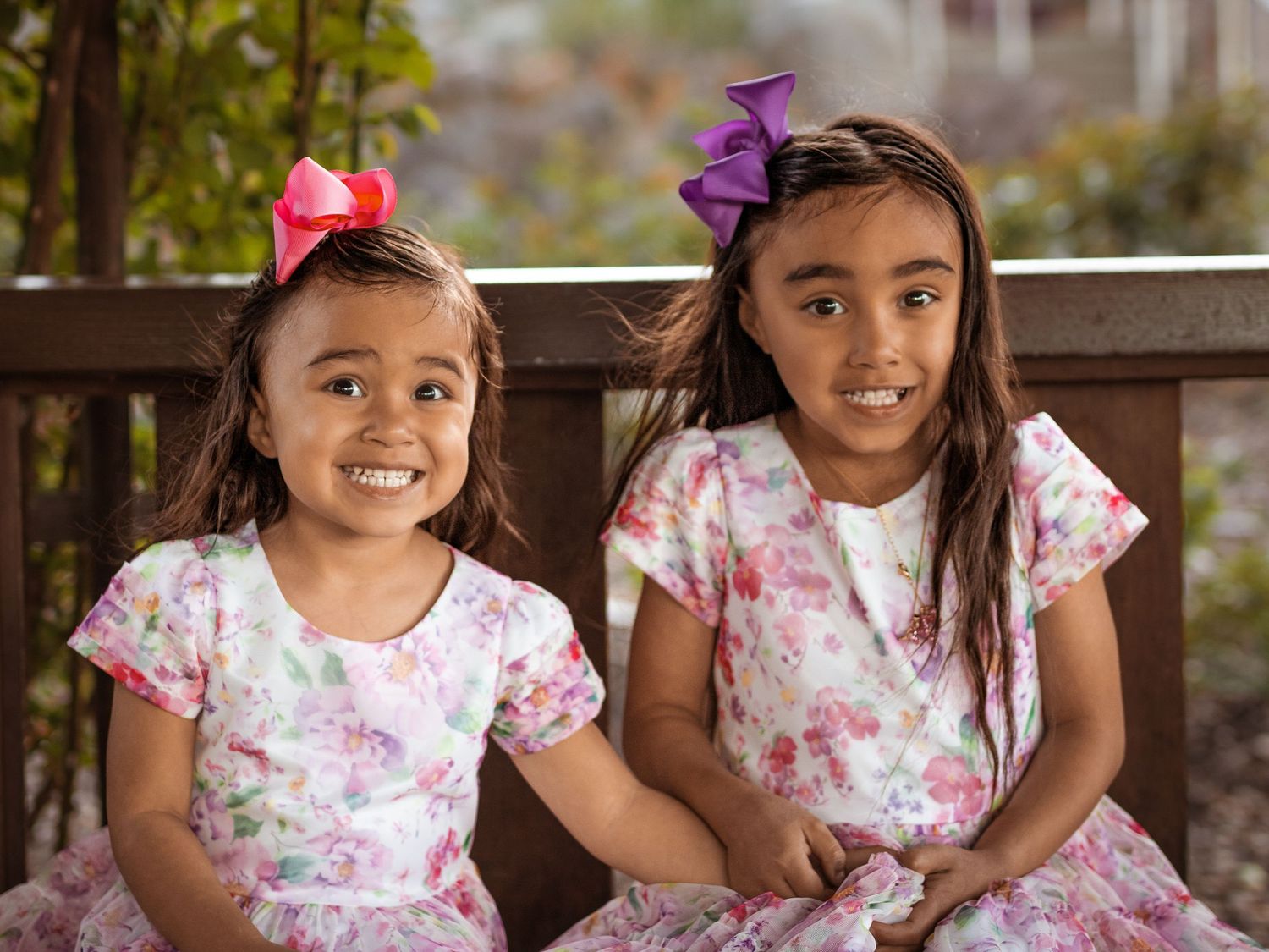 Two young girls in matching floral dresses and hair bows smile brightly for the camera.