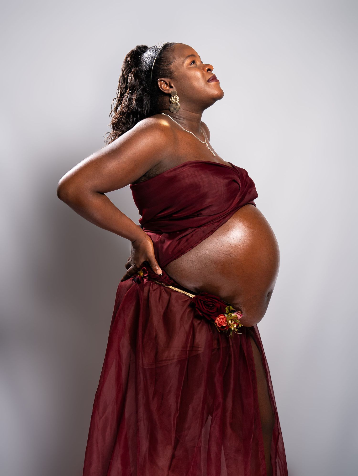 Woman in burgundy dress poses against white background in artistic maternity photoshoot.