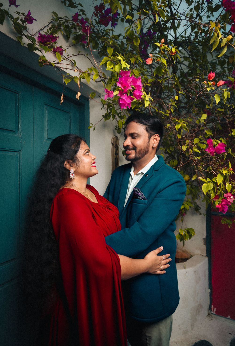 A couple poses by a teal door with pink bougainvillea flowers overhead.