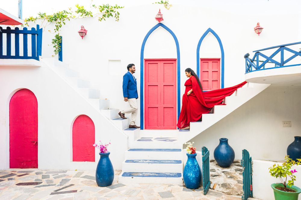 A couple poses on white stairs with red doors and blue arches in a Mediterranean setting.