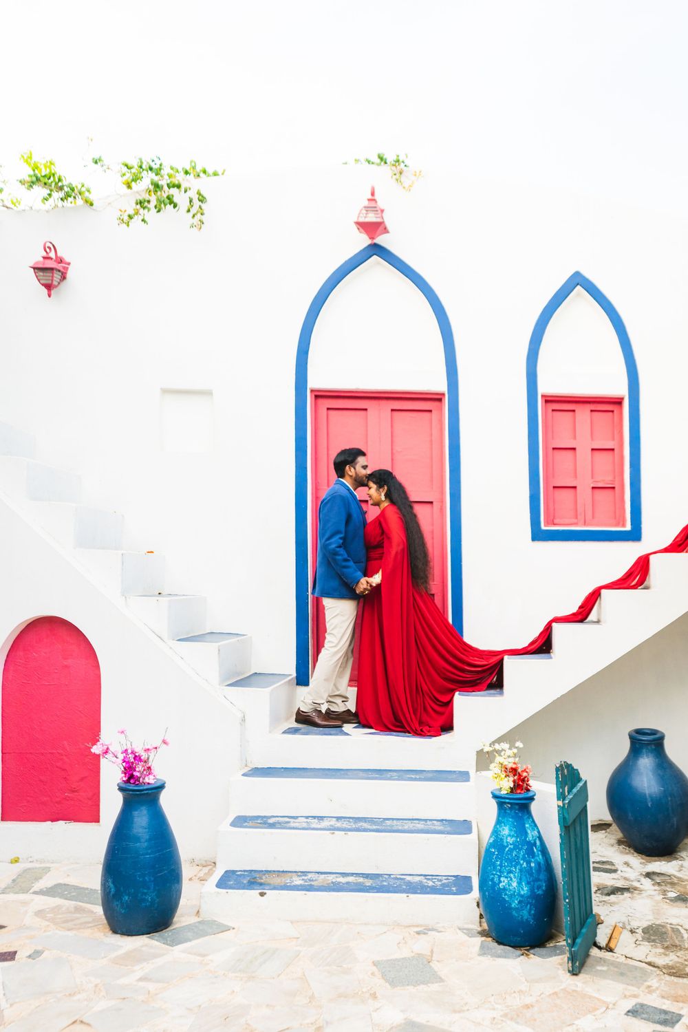 A couple embraces on white steps with red doors and blue architectural details.