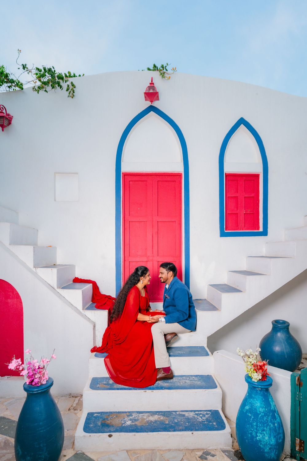 A couple in red and blue outfits sit on white Mediterranean-style steps with arched doorways.