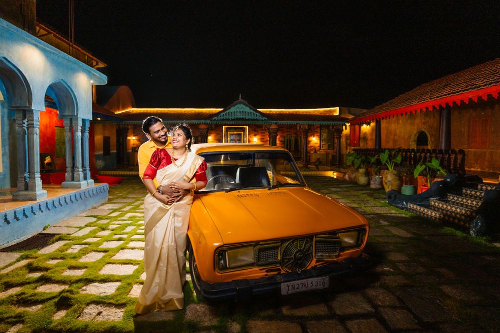 A couple embraces near a vintage car at night with blue-lit architecture in background.