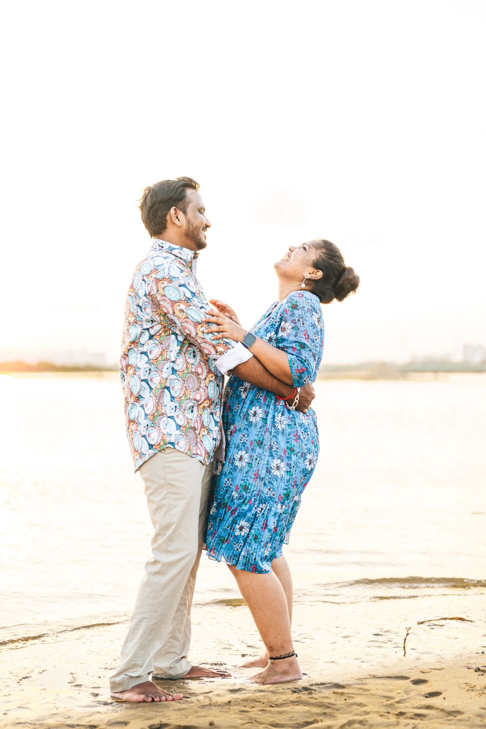 A couple in floral print clothing laughing together on a sandy beach at sunset.