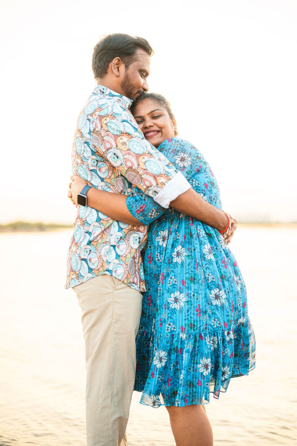 A couple in matching floral outfits embrace while standing on a sandy beach at sunset.