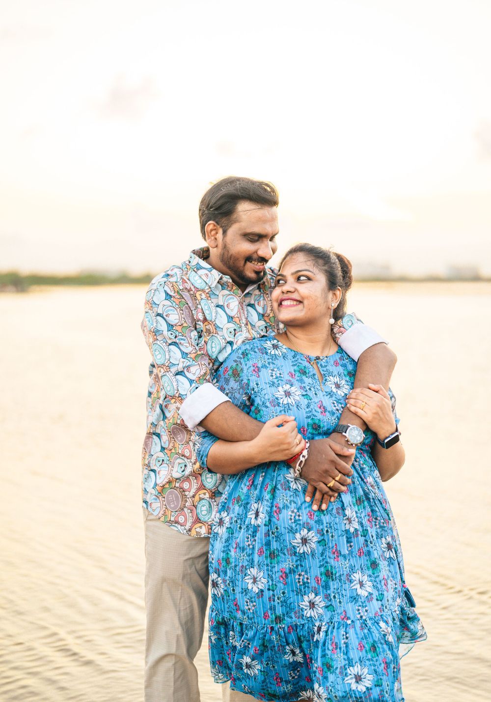 A couple in coordinating blue and floral outfits embraces on a beach during golden hour.