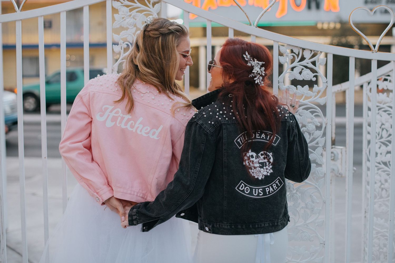 Two people wearing custom jackets pose back-to-back against a white fence.