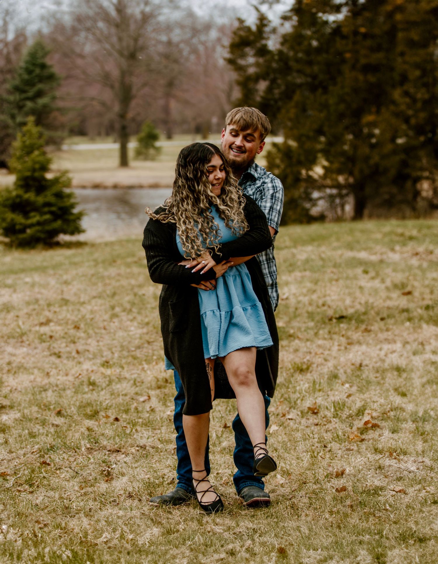 A couple sharing an intimate embrace on grassy terrain with evergreen trees in the background.