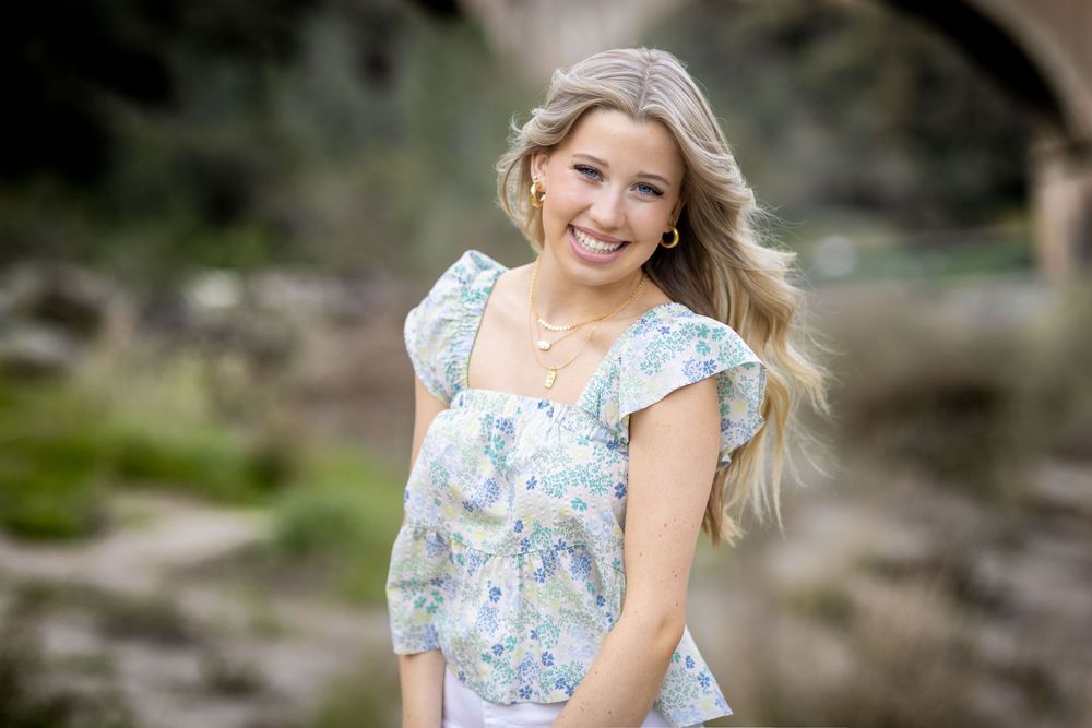 A woman in a light blue floral dress smiles while posing outdoors in a natural setting.