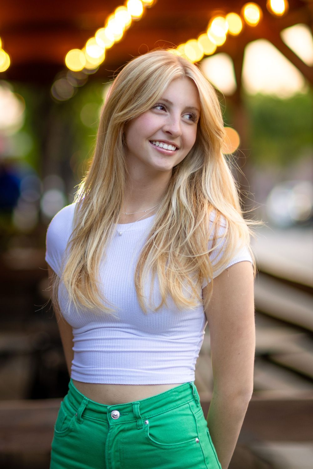 Person in white top and green pants smiling against bokeh lights background.