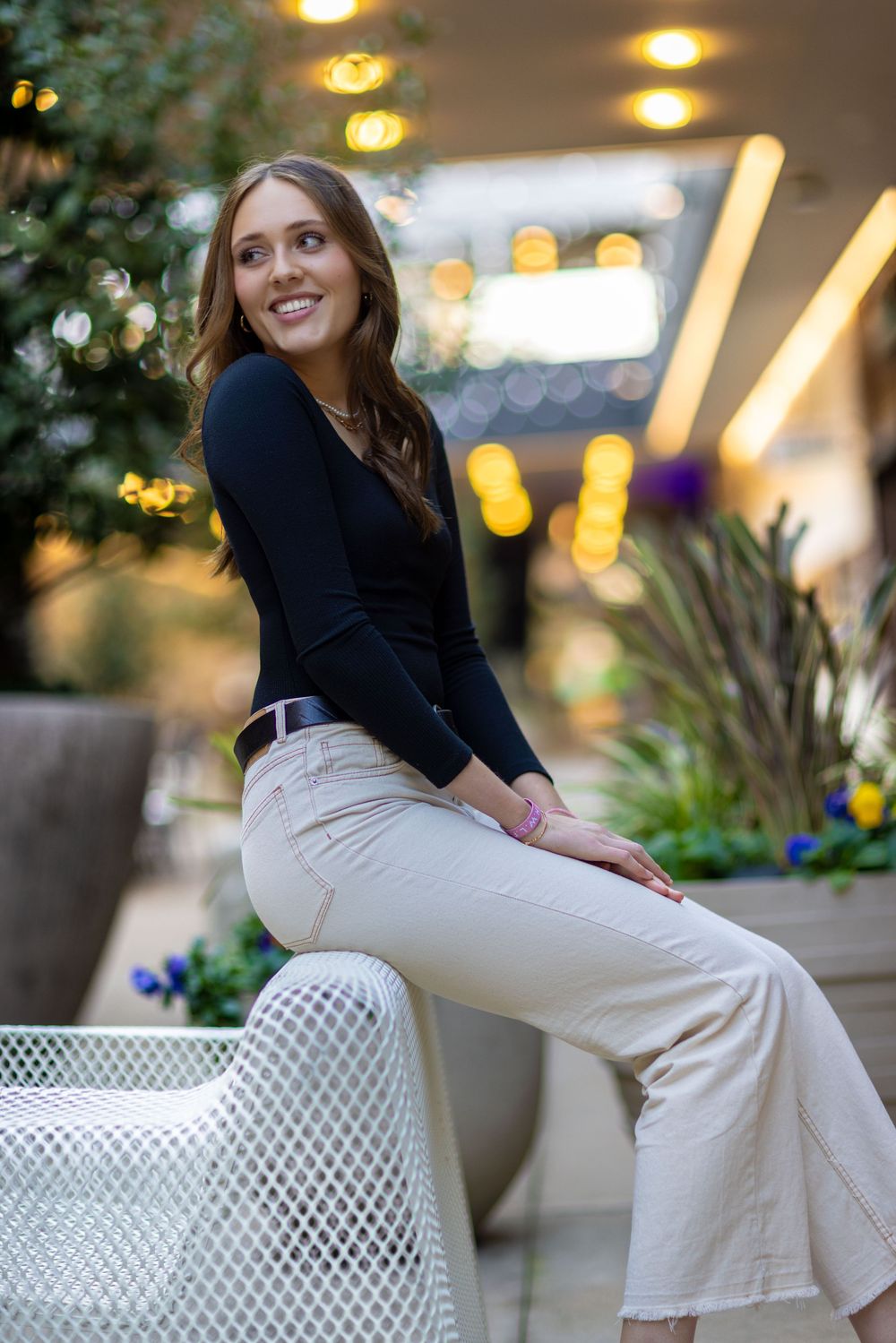 Person in black top and light pants sits casually on white outdoor furniture with bokeh lighting in background.