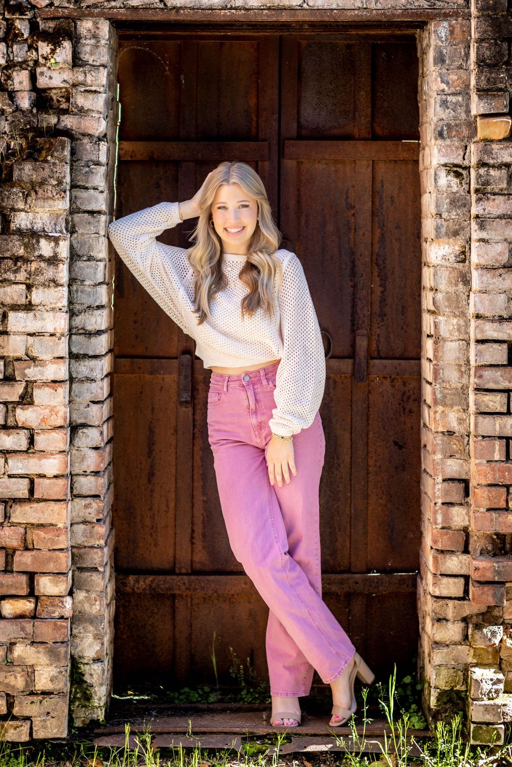 A person in pink pants and white top poses playfully against rustic brick archways in an outdoor setting.