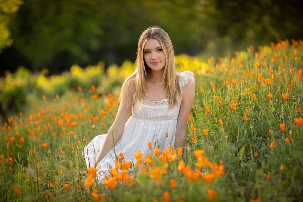 A person in a white dress sits among vibrant orange wildflowers in a sunny meadow during golden hour.