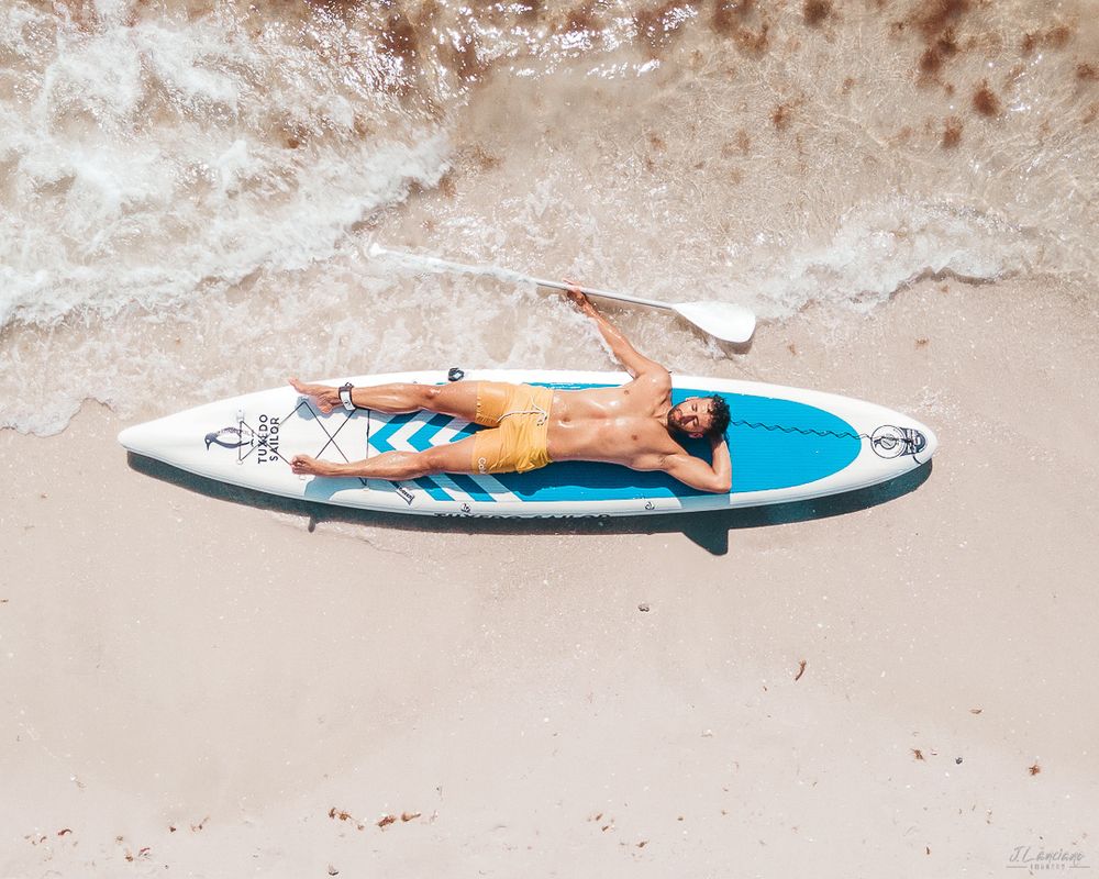 Aerial view of person relaxing on paddleboard on sandy beach.
