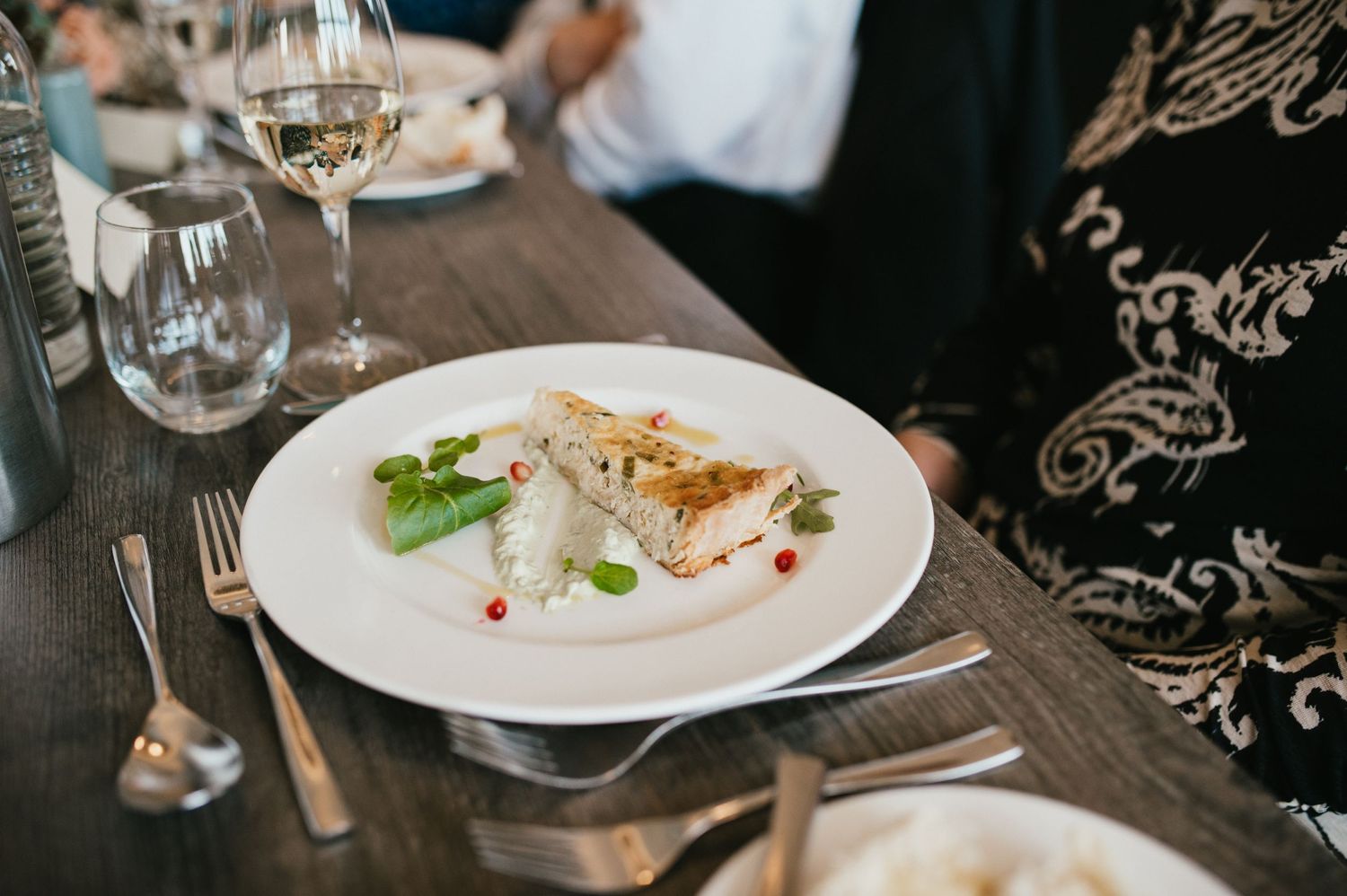 Elegant plated dish with garnishes and white wine glass on dark wooden table.