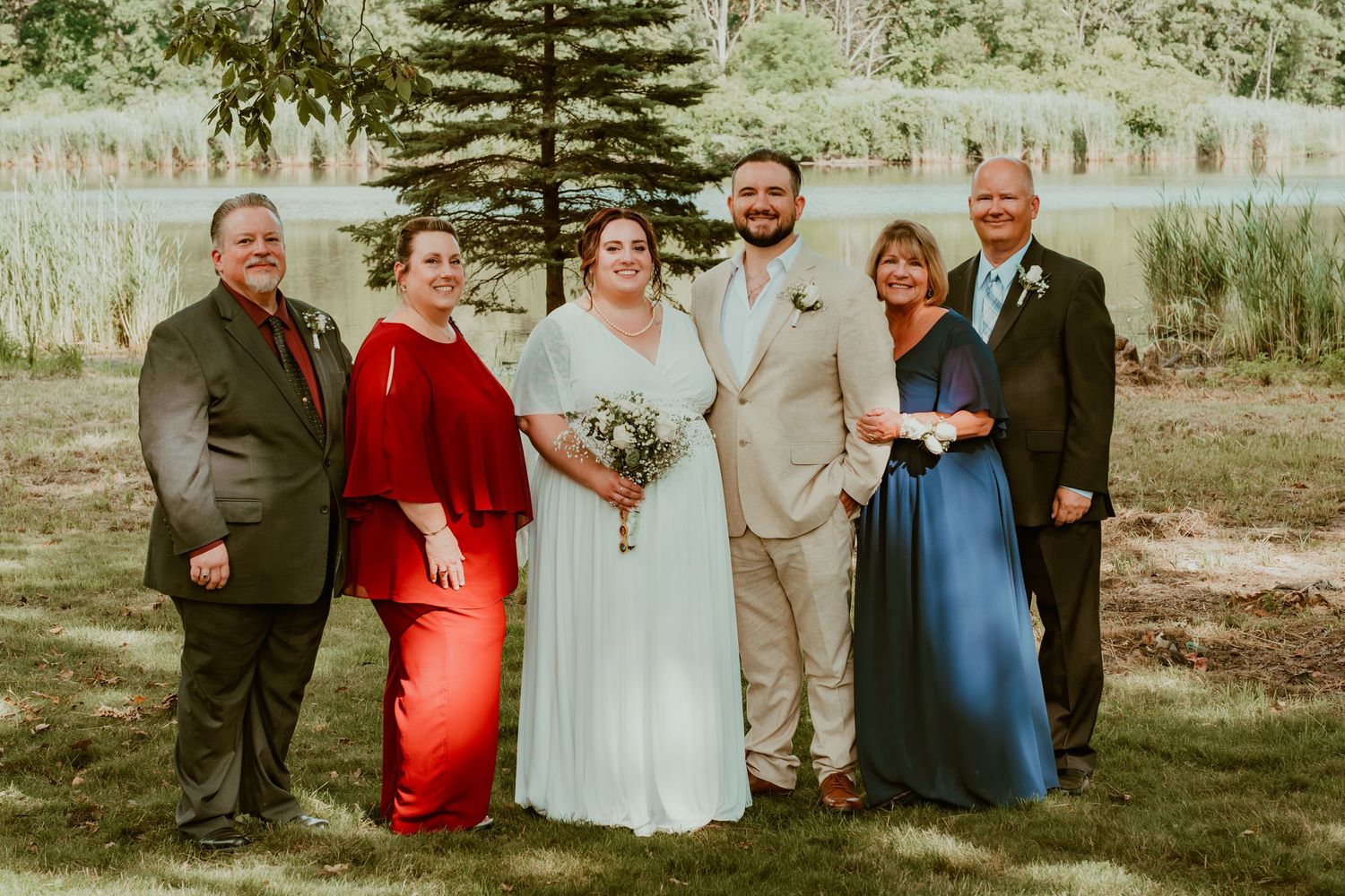 A wedding group poses together outdoors near a serene lake and pine trees.