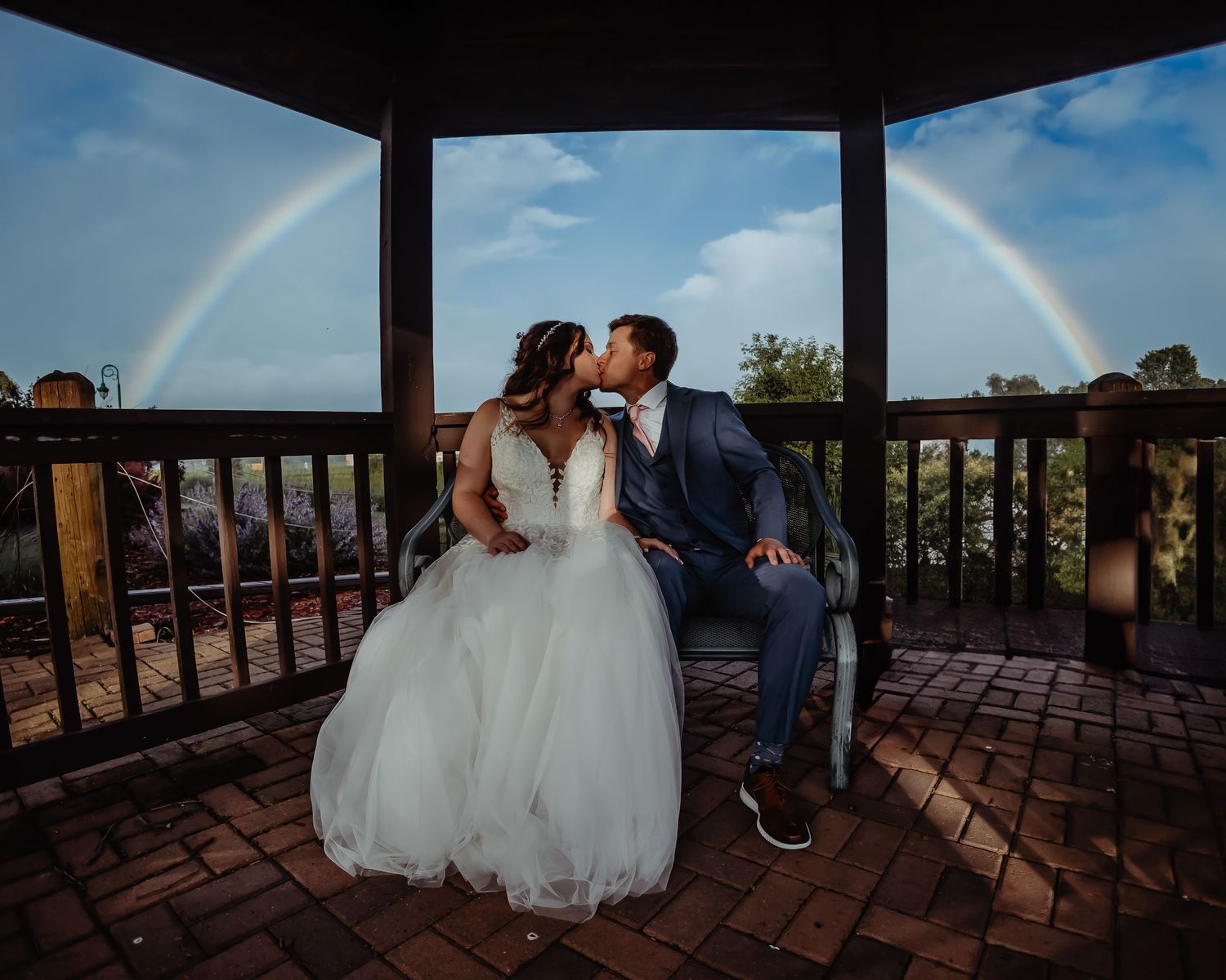 Wedding couple under gazebo with perfect double rainbow arching across blue sky.