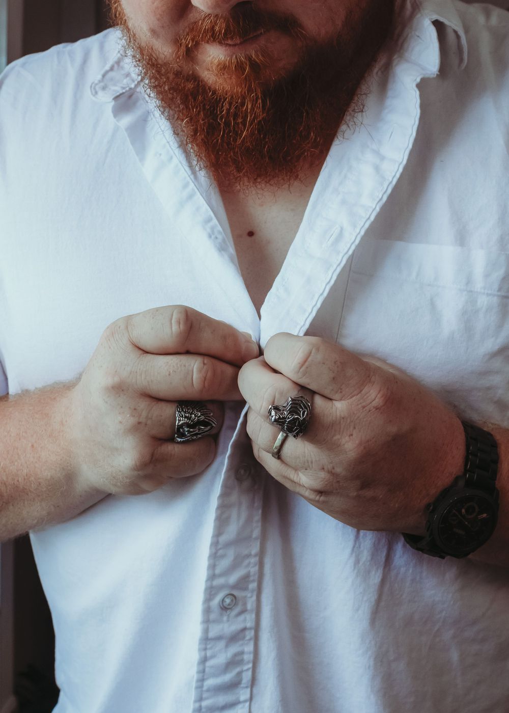 Close-up of hands buttoning white dress shirt with visible rings and ginger beard.