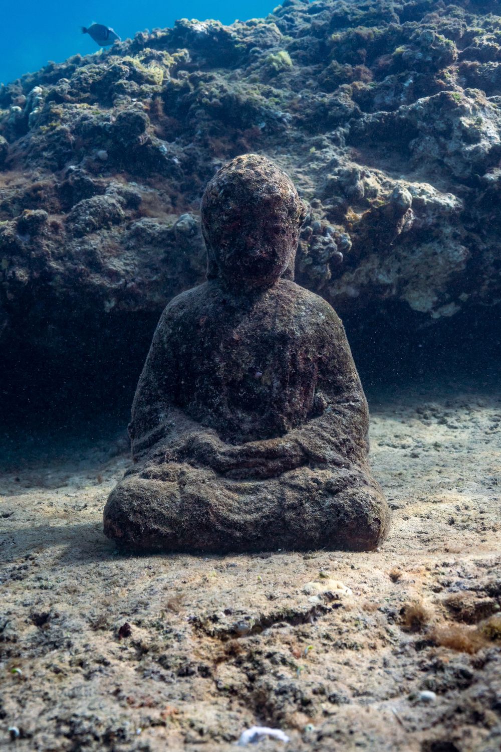 Ancient stone Buddha statue sitting on ocean floor surrounded by coral reef formations.