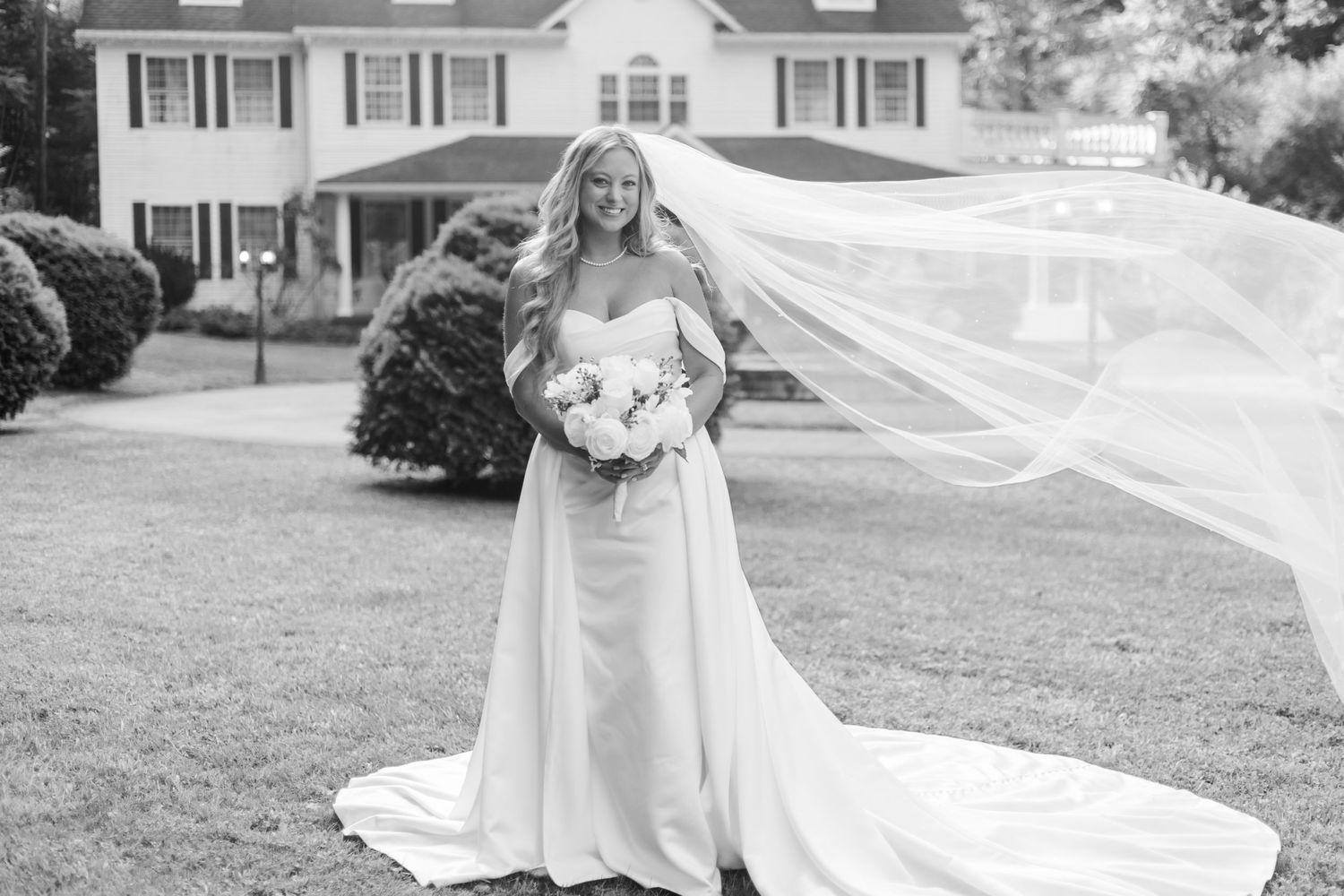 Bride's veil flows dramatically in the wind against classic white house backdrop.
