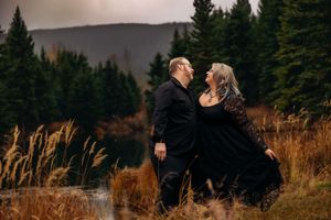 A romantic couple in black outfits embrace against a moody mountain landscape with evergreen trees at sunset.