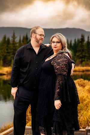Couple in coordinating black formal wear pose together near a lake with mountains in background.